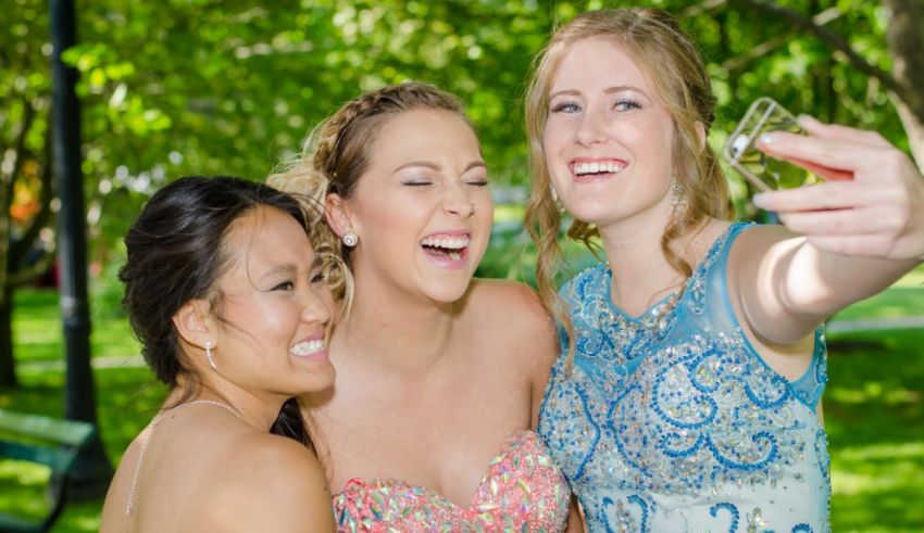 Three girls in formal dresses taking a selfie in a park.