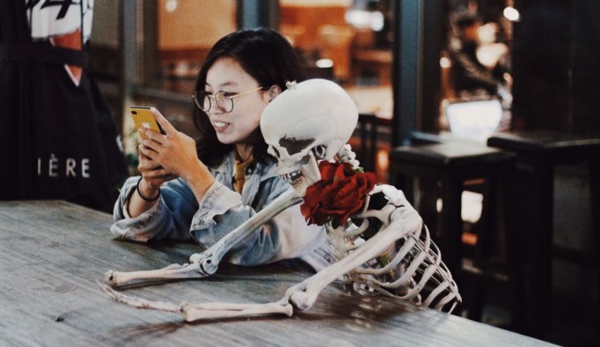 A woman sitting at a table with a skeleton and a cell phone.