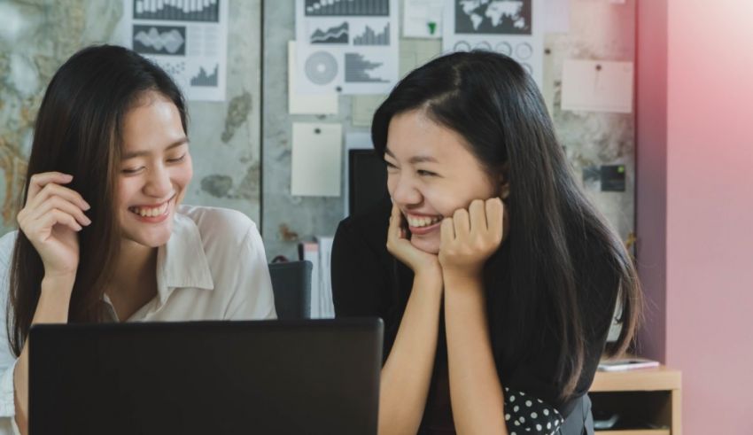 Two asian women smiling at each other while working on a laptop.