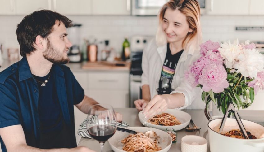 A man and a woman eating spaghetti in a kitchen.
