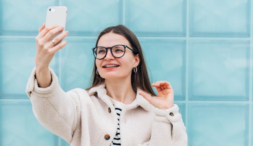 A woman in glasses is taking a selfie in front of a blue wall.