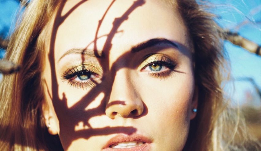 A woman with blond hair and blue eyes is posing in front of a tree.