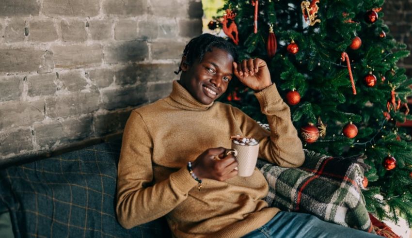 A black man sitting on a couch near a christmas tree with a cup of coffee.