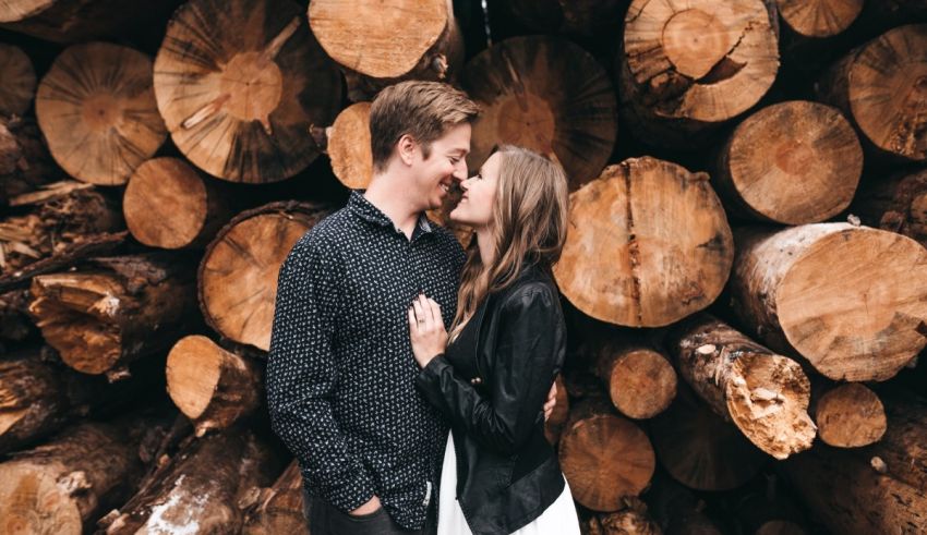 A couple kisses in front of a pile of logs.