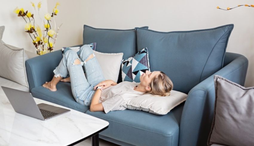 A woman is laying on a blue couch with her laptop.