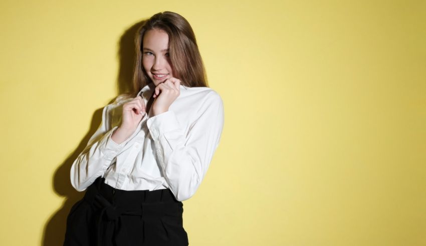 A young woman in a white shirt and black pants posing against a yellow background.