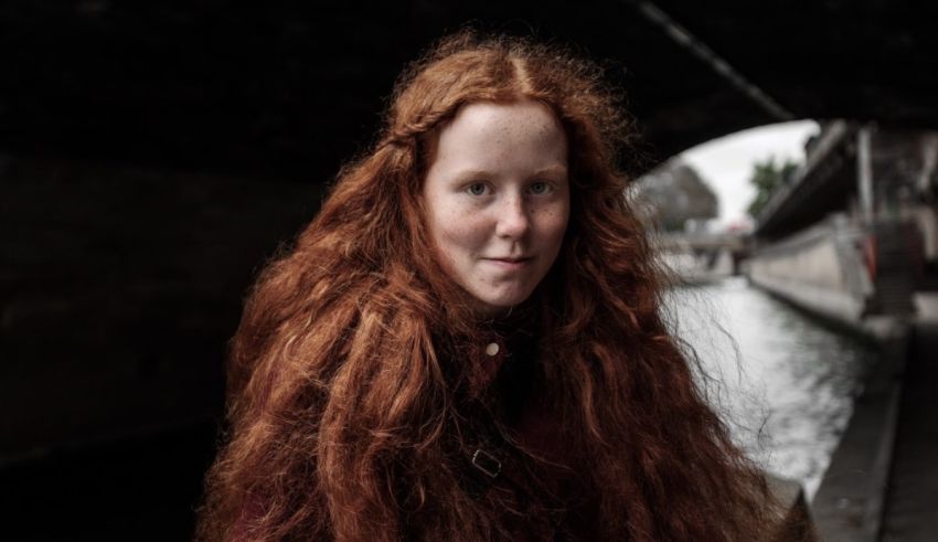 A girl with long red hair is standing under a bridge.