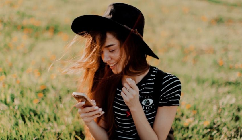 A girl in a hat sitting in a field looking at her phone.