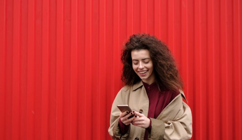 A woman with curly hair is using her phone in front of a red wall.