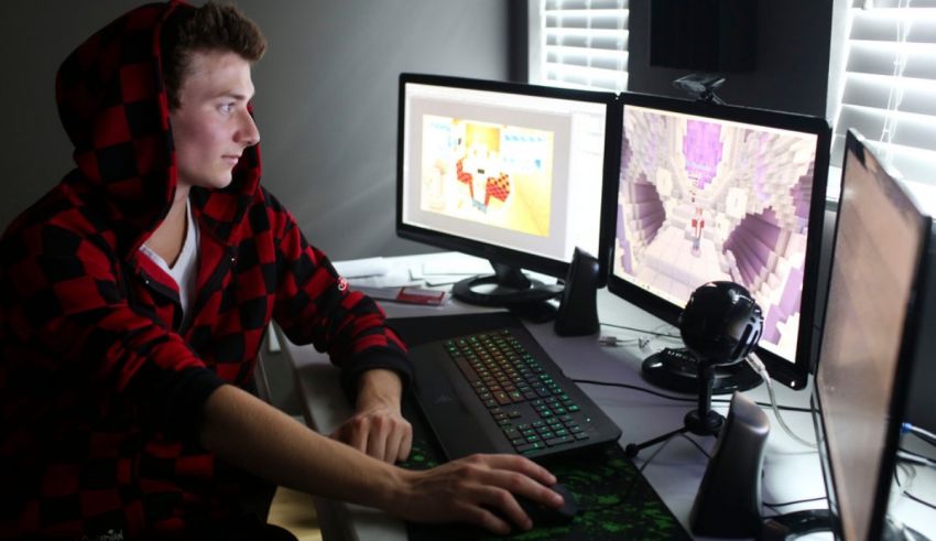 A young man in a red hoodie sitting at a desk with three monitors.