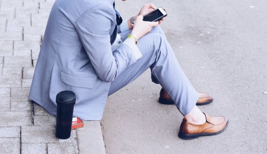 A man in a suit is sitting on the sidewalk and using his cell phone.