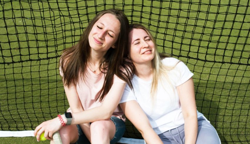 Two girls sitting on the ground next to a tennis net.