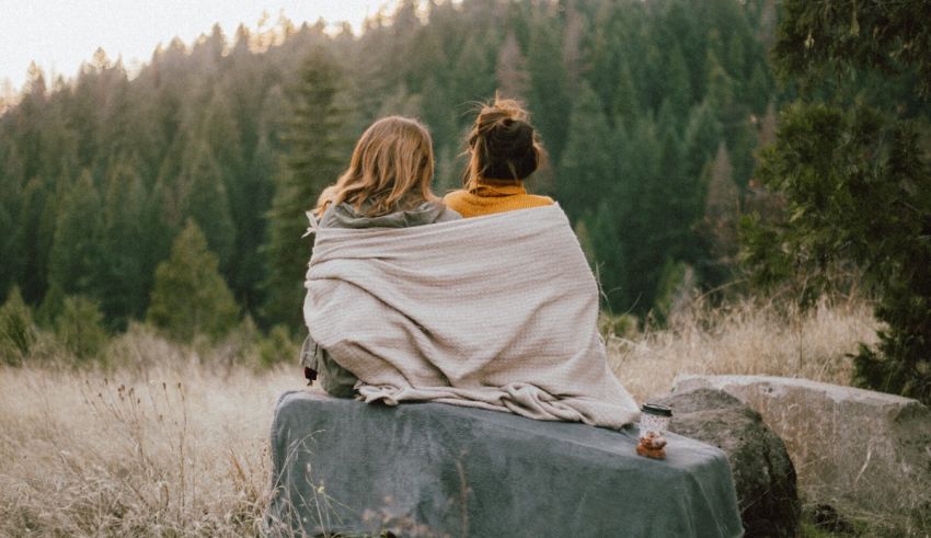 Two women wrapped in blankets sitting on a rock in a forest.
