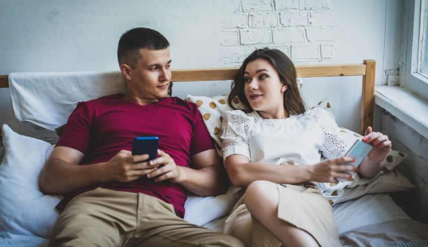 A young man and woman are sitting on a bed looking at their cell phones.