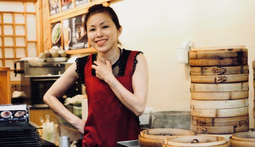 A woman in a red apron standing in front of a stack of dumplings.