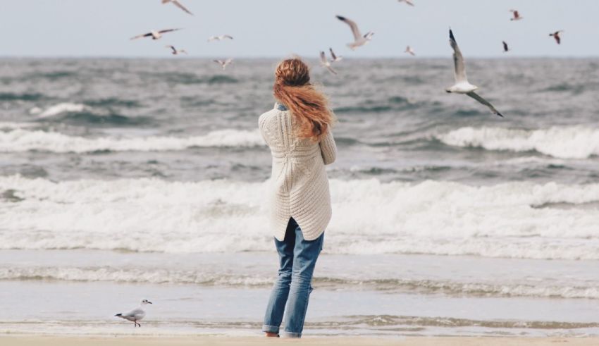 A woman is standing on the beach with seagulls flying around her.