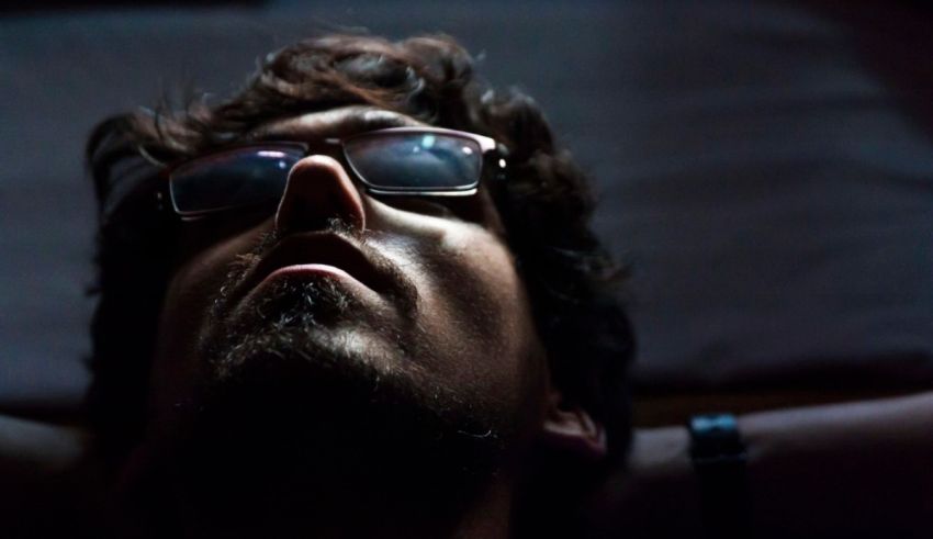 A man with glasses laying down in the dark.