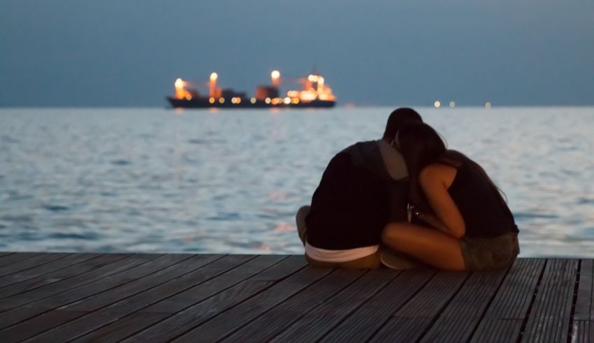 Two people sitting on a dock with a ship in the background.