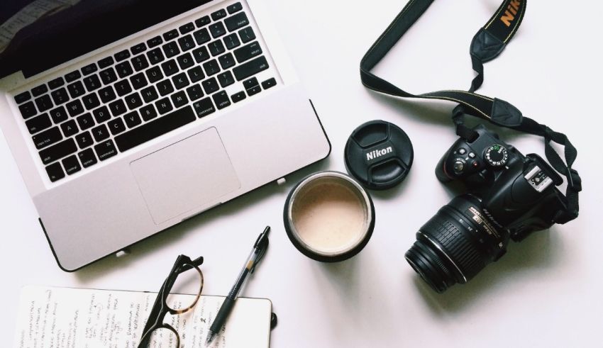 A laptop, a camera, a notebook and a cup of coffee.