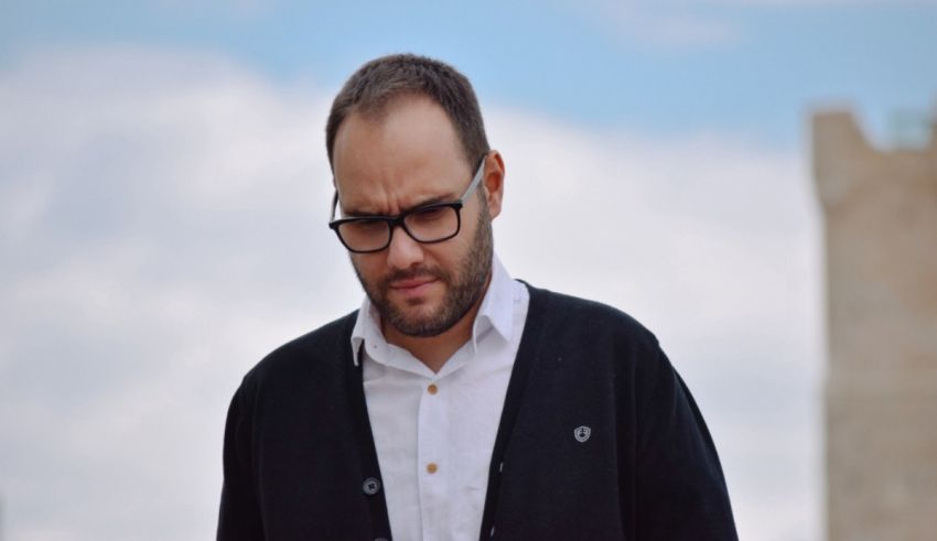 A man wearing glasses and a cardigan is standing in front of a castle.