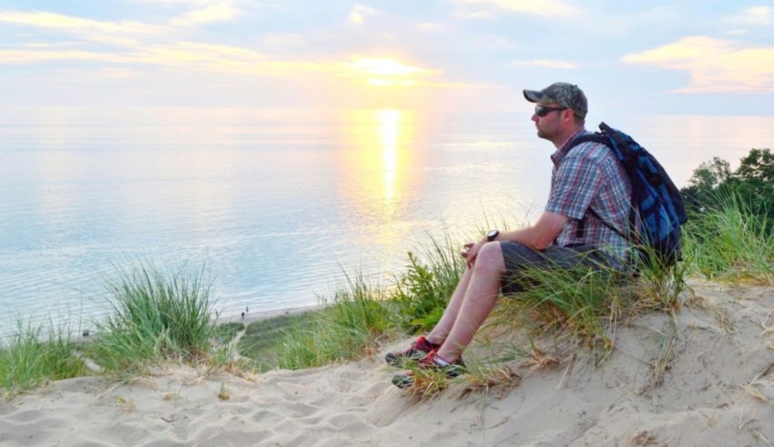 A man sits on top of a sand dune at sunset.