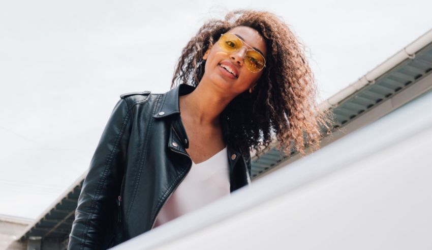 A young woman wearing sunglasses and a leather jacket leaning against a car.