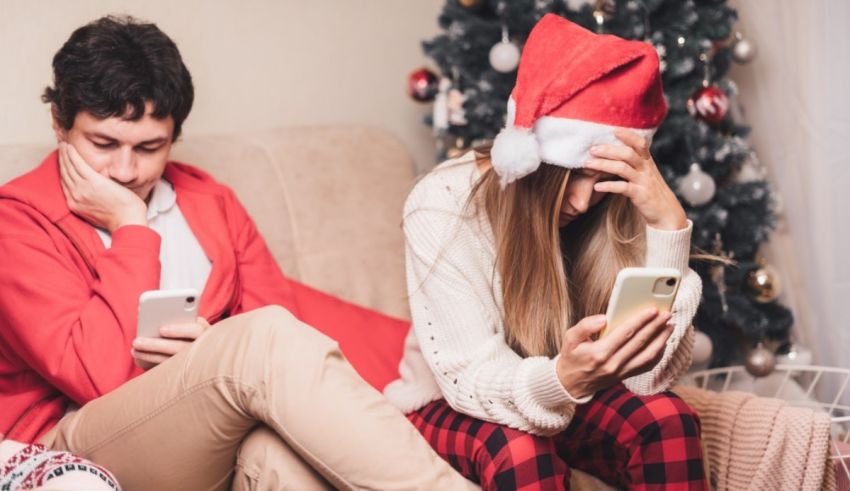 A man and woman sitting on a couch with santa hats looking at their phones.