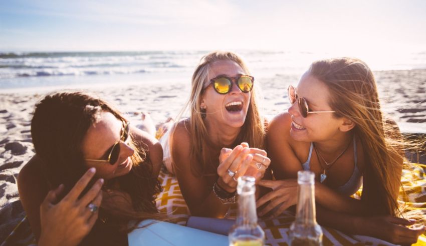 Three women laughing on the beach with a bottle of beer.