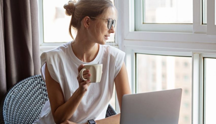 A woman sitting in front of a window with a laptop and a cup of coffee.