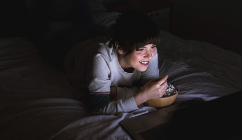 A woman laying on a bed with a bowl of popcorn and a laptop.