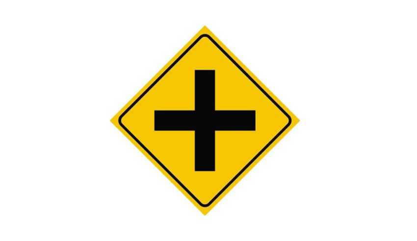 A yellow road sign with a black cross on it.