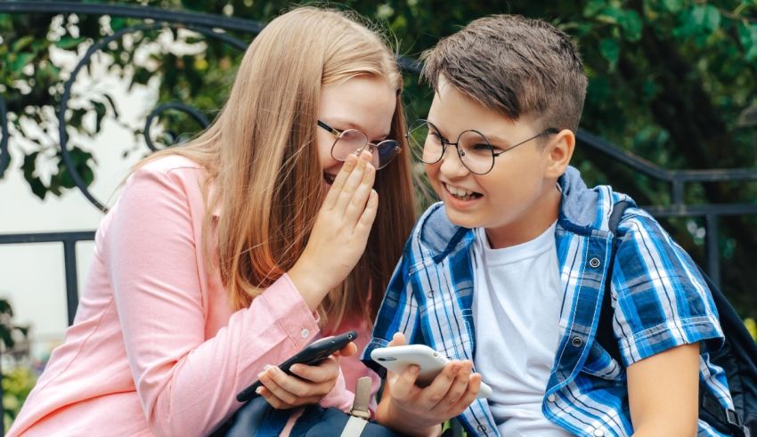 A boy and girl are sitting on a bench and looking at their cell phones.