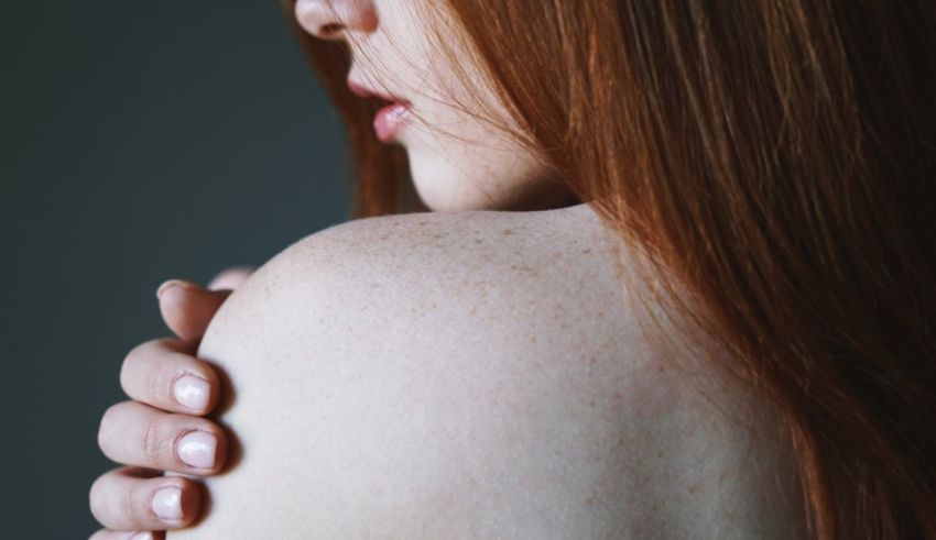 A woman with red hair is posing with her hand on her shoulder.