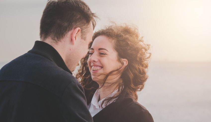 A man and woman are smiling at each other in front of the ocean.