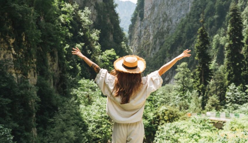 A woman in a hat standing on a path in the mountains with her arms outstretched.