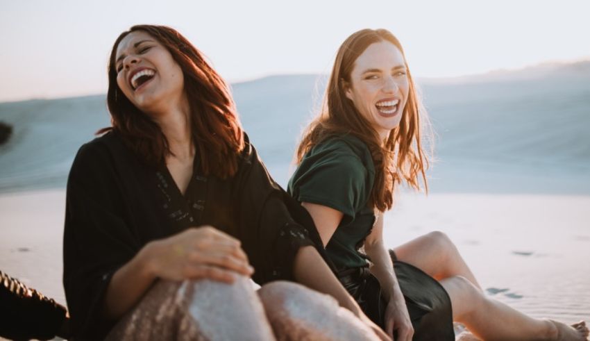Two women laughing in the sand at sunset.