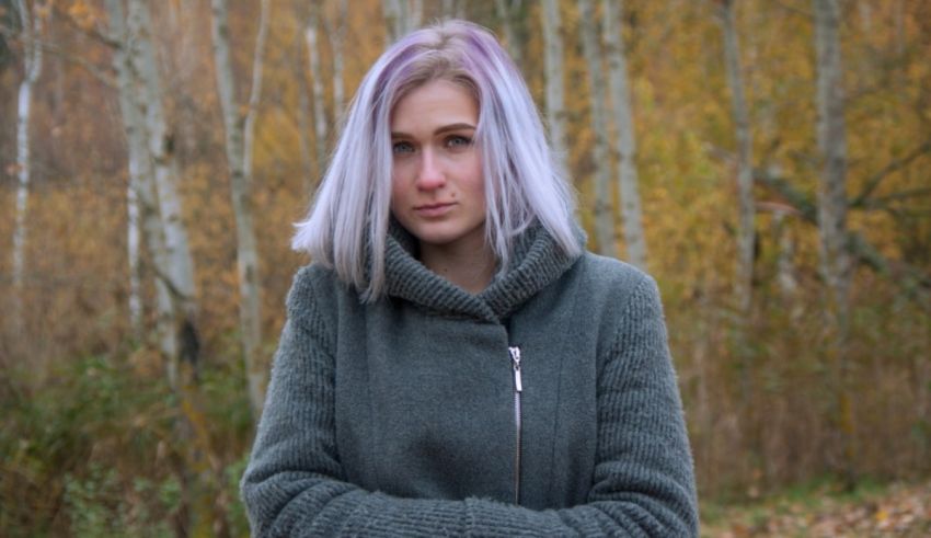 A young woman with purple hair standing in a wooded area.