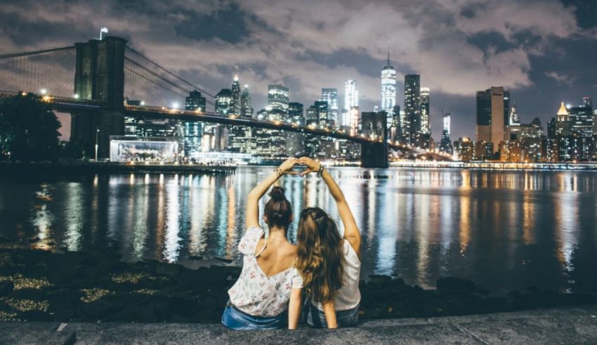 Two young women making a heart shape in front of the brooklyn bridge at night.