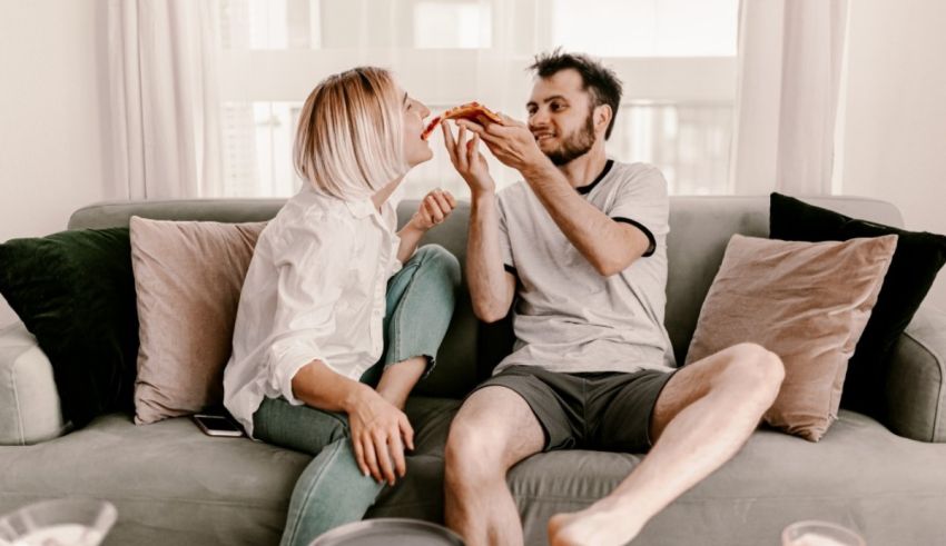 A man and woman sitting on a couch eating pizza.