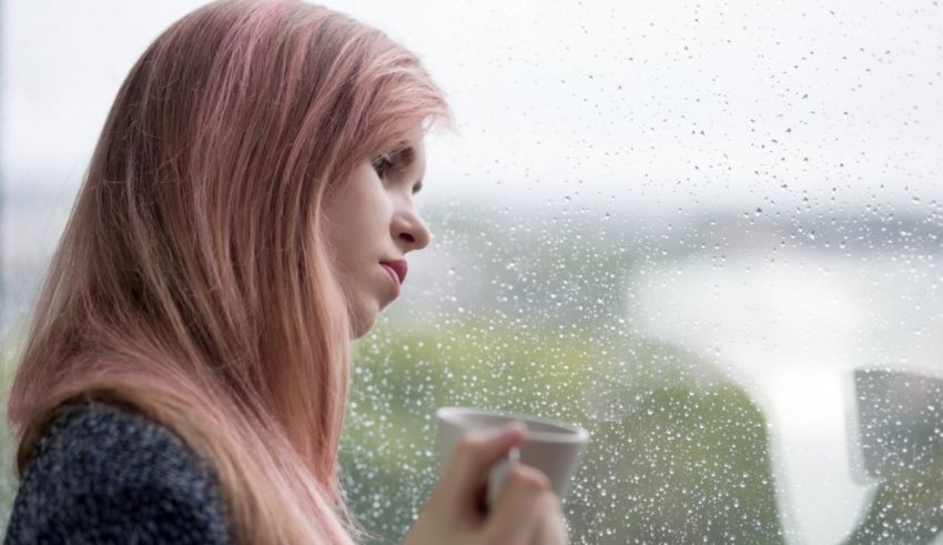 A girl with pink hair looking out the window with a cup of coffee.