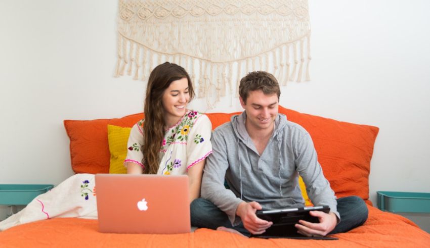 A man and woman sitting on a bed using a laptop.