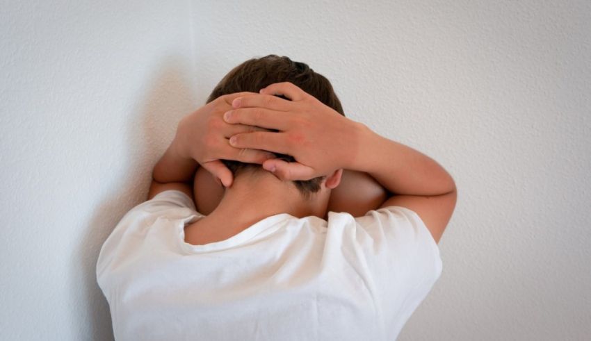A boy covering his head with his hands in front of a white wall.