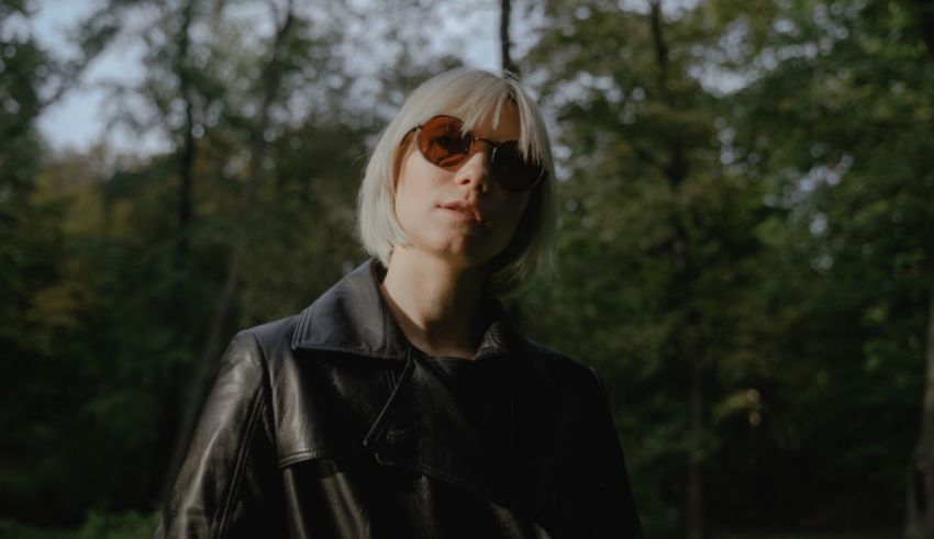 A woman in a leather jacket and sunglasses is standing in a wooded area.
