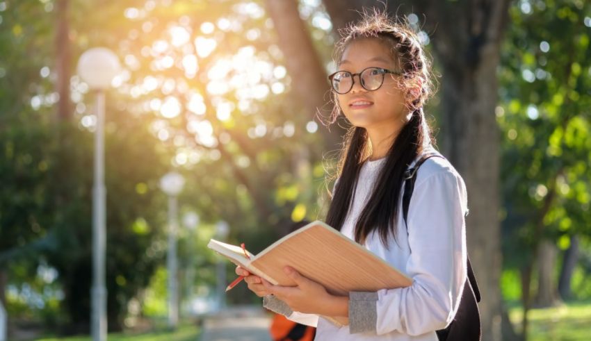 A young asian girl wearing glasses and holding a book in the park.