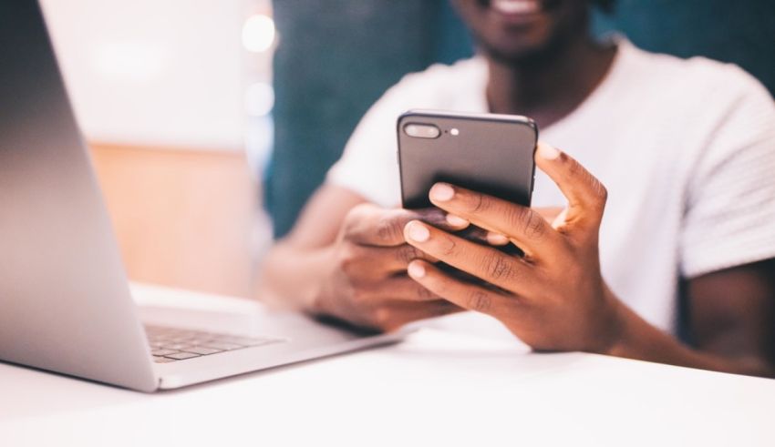 A man is using a smartphone while sitting in front of a laptop.