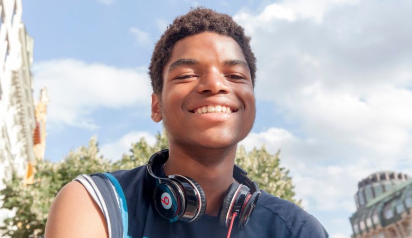 A young man wearing headphones smiles in front of a city.