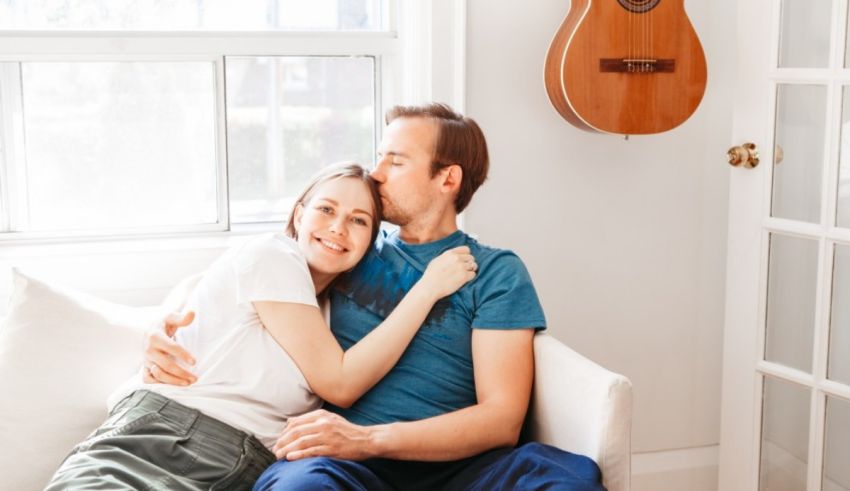 A couple sitting on a couch with an acoustic guitar in front of a window.