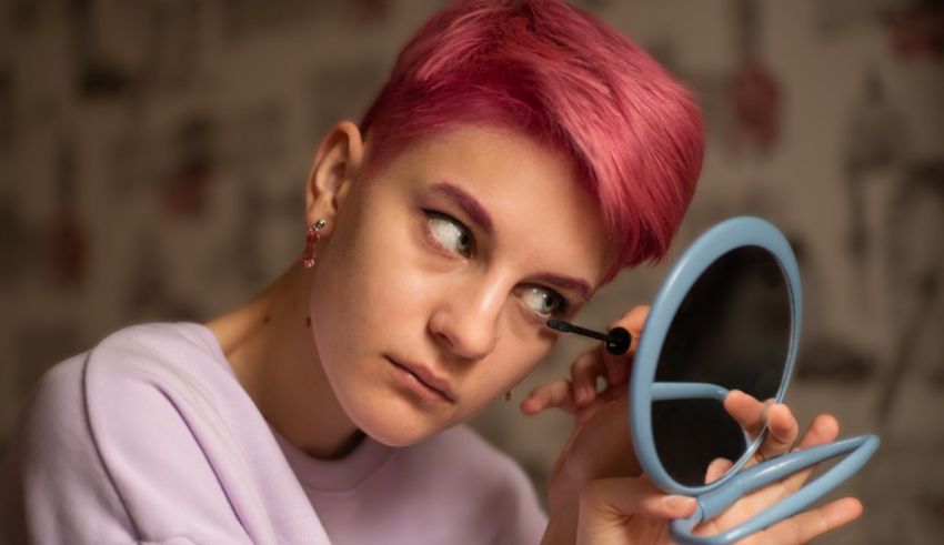 A woman with pink hair is putting makeup on with a mirror.