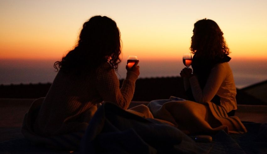 Two women sitting on a blanket with wine glasses at sunset.