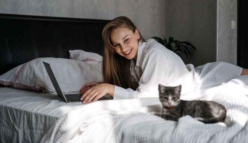 A woman is sitting on a bed with a cat next to her laptop.